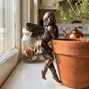 May include: A bronze-toned fairy figurine with detailed wings, a short dress, and boots, designed to hang over the edge of a terracotta flower pot. The figure has wavy hair and is leaning its head on one hand while perched on the rim.