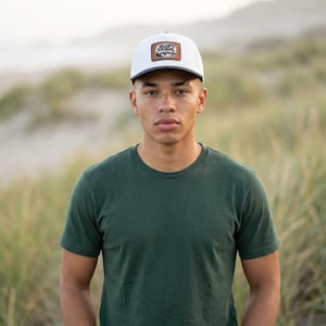 May include: A white baseball cap with a dark brim and a rectangular brown patch on the front featuring the text Old Tahoe above a mountain and forest graphic, paired with a solid dark green short-sleeved crew neck t-shirt.