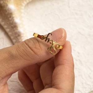 May include: A gold-toned metal ring featuring a cursive script design that spells out the name Skye. The ring has a thin, polished band and the lettering is integrated into the top section of the piece, creating a continuous, flowing aesthetic.