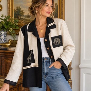 Könnte beinhalten: Eine langärmelige Jacke mit Knopfleiste und einem Color-Block-Design aus cremefarbenen und schwarzen Bahnen. Die Jacke verfügt über zwei Vordertaschen, eine mit einem perlenbesetzten Giraffenmotiv und die andere mit einem Elefantenmotiv sowie Spitzenbesatz.