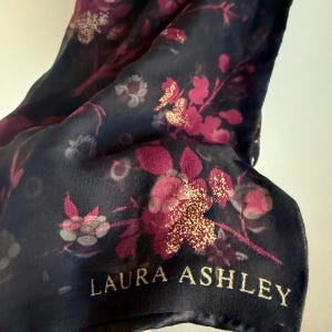 May include: A dark navy blue scarf featuring a floral pattern in shades of magenta and deep pink, with gold glitter accents on the petals. The fabric is semi-sheer and lightweight. The text LAURA ASHLEY is printed in gold lettering at the bottom.
