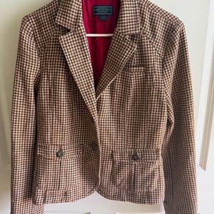 May include: A brown and white houndstooth blazer featuring a notched lapel, long sleeves, and two front flap pockets with buttons. The interior is lined in solid red fabric with an American Eagle Outfitters label at the neck.