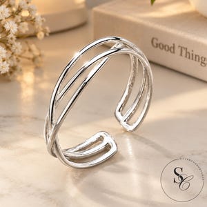 Puede incluir: Un brazalete tipo puño de tono plateado pulido con un diseño de múltiples hilos con tres bandas superpuestas que convergen en los extremos. El brazalete abierto tiene una superficie lisa y reflectante e incluye un logotipo circular con el texto SILVERCHARMBYSAAD.