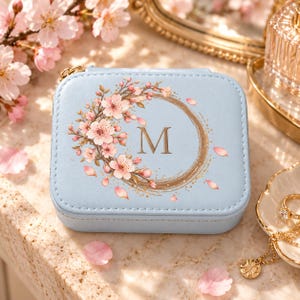 Puede incluir: Un joyero cuadrado de color azul claro con una letra M dorada centrada dentro de una corona circular de flores de cerezo rosas. La caja tiene un exterior liso y estructurado con un tirador de cremallera de color dorado.