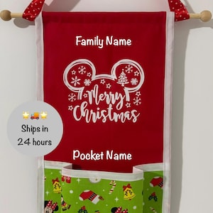 May include: A red fabric wall hanging with a wooden dowel and a red polka dot ribbon for suspension. The red section features white text reading Family Name, a white Mickey Mouse ear outline with snowflakes, and the text Merry Christmas. Below is a green pocket with a repeating pattern of Minnie Mouse, snowmen, candy canes, and bells, labelled with the text Pocket Name.