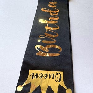 May include: A black fabric sash featuring the text Birthday Queen in a reflective gold metallic font. Below the text is a graphic of a gold metallic crown with the word queen written in a black script font across the centre of the crown.