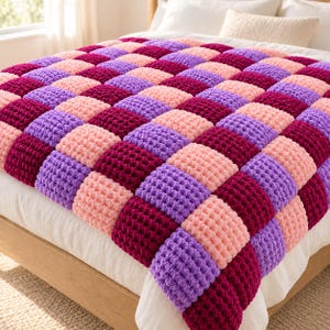 Puede incluir: Una manta de punto grueso con un patrón de tablero de ajedrez repetitivo de bloques cuadrados en tonos magenta intenso, lavanda claro y rosa pastel suave. La textura es gruesa y afelpada, con cada cuadrado mostrando un punto de bucle constante.