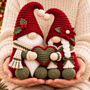 May include: Two crocheted gnomes with red pointed hats, one featuring holly leaves and the other a poinsettia. They have beige faces, white beards and braids, and olive green feet. The gnomes hold a dark green heart between them.