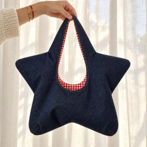 May include: A star-shaped handbag crafted from dark blue denim fabric. The interior lining features a red and white gingham check pattern. The bag has a single integrated handle opening at the top and visible stitching along the edges.
