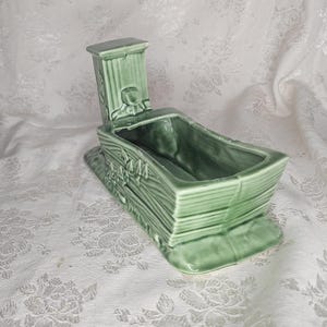 May include: A green ceramic planter shaped like a boat with a hinged lid. The planter has a rectangular base and is decorated with a wood grain texture and floral accents. The lid has a small figure on it.