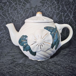May include: White ceramic teapot with a green and gold floral design. The teapot has a rounded body and a lid with a gold knob.