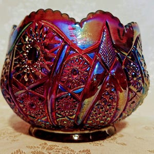 May include: A decorative, iridescent glass bowl with a scalloped rim. The bowl features a deep red color with blue and gold highlights. The exterior is patterned with geometric and floral designs, and it sits on a dark brown base.