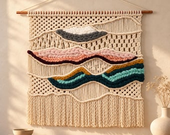 May include: A wall hanging made of cream-colored macrame cord suspended from a wooden rod. The piece features woven sections of thick yarn in shades of gray, pink, burgundy, mustard yellow, teal, and light blue, finished with a long fringe.