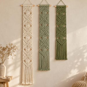 May include: Three vertical macrame wall hangings suspended from wooden dowels. The left piece is cream, the center is sage green, and the right is olive green. Each features a repeating diamond pattern with intricate knotted cord details.
