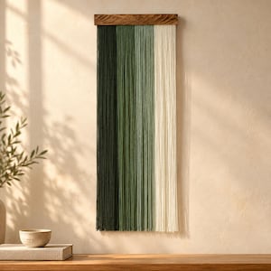 May include: A wall hanging featuring vertical strands of fiber in an ombre gradient, transitioning from dark forest green to sage green and ending in cream. The strands are suspended from a horizontal, rectangular wooden bar.