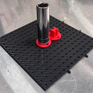 May include: A square black pegboard with a carbon fibre texture featuring a grid of holes. A tall, cylindrical chrome metal socket is mounted on a red circular base, and a separate red H-shaped plastic component sits nearby on the board.