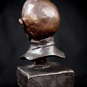 May include: Bronze bust sculpture of a person's head and shoulders. The sculpture is on a square base. The bust is facing to the left, but the head is turned to the right. The sculpture is a dark brown color.