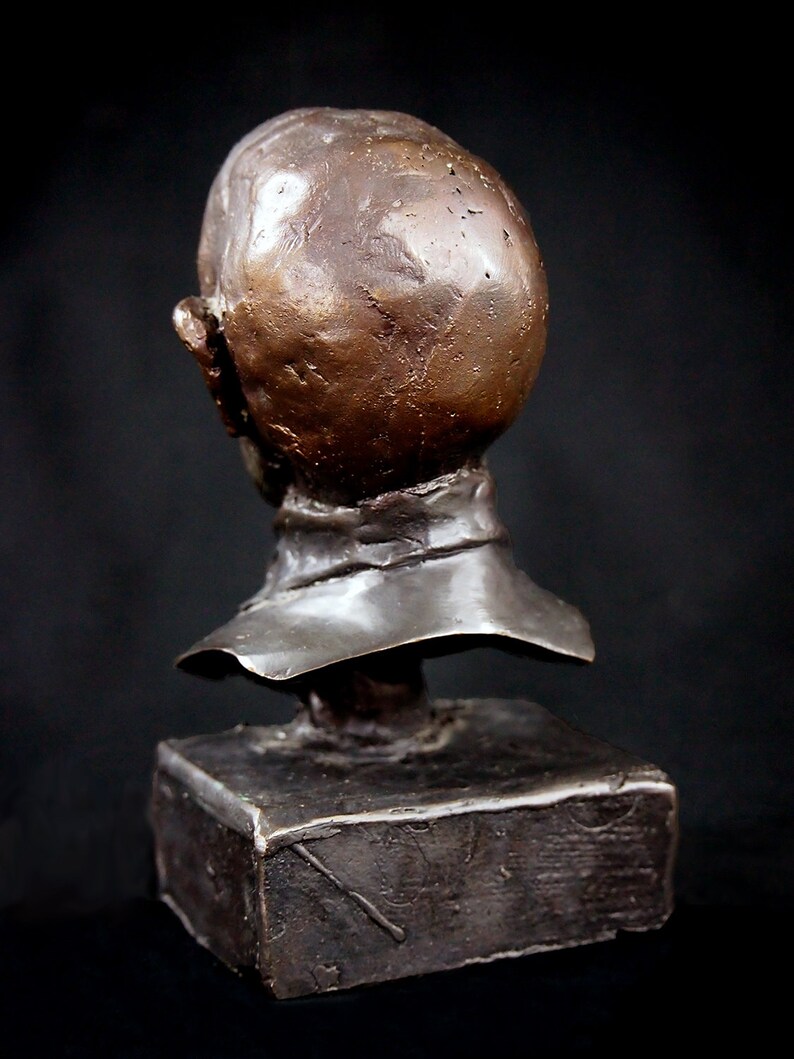 May include: Bronze bust sculpture of a person's head and shoulders. The sculpture is on a square base. The bust is facing to the left, but the head is turned to the right. The sculpture is a dark brown color.