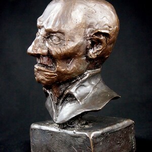 May include: Bronze bust sculpture of a man's head and shoulders. The sculpture is on a square base. The man has a wrinkled face and a serious expression.