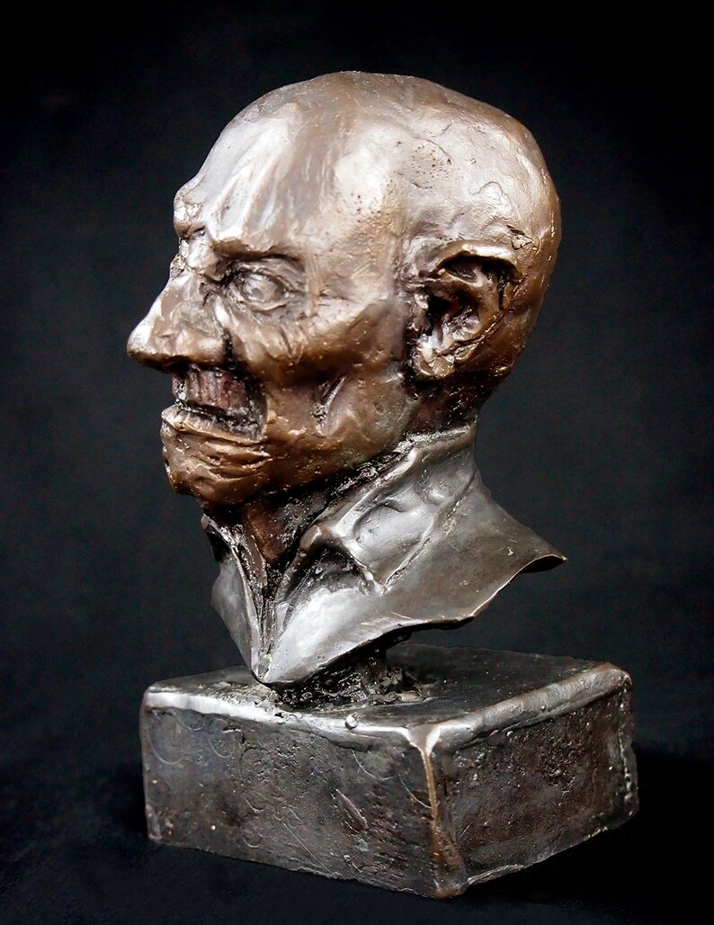 May include: Bronze bust sculpture of a man's head and shoulders. The sculpture is on a square base. The man has a wrinkled face and a serious expression.