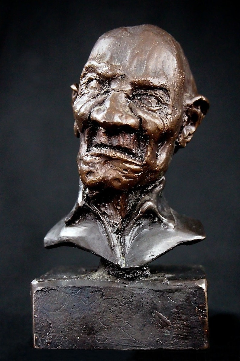 May include: Bronze bust sculpture of a man with a wrinkled face and a mischievous expression. The sculpture is mounted on a square base.