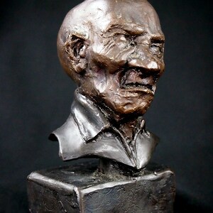 May include: Bronze bust sculpture of a man with a weathered face and a furrowed brow. The sculpture is mounted on a dark brown rectangular base.