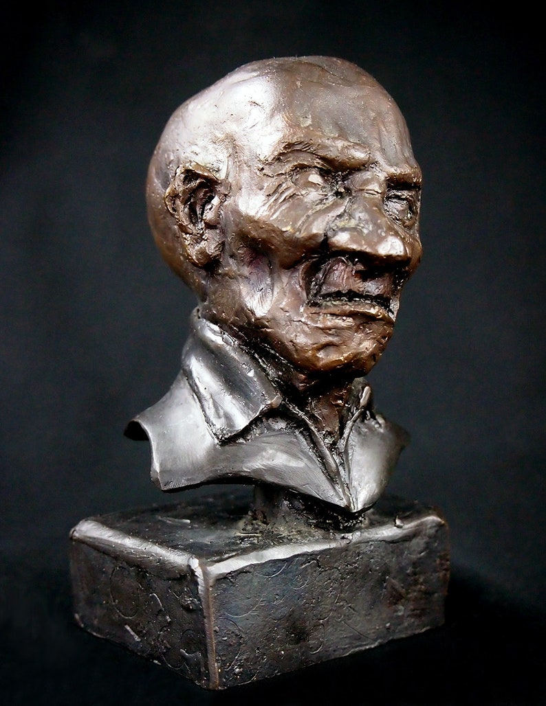 May include: Bronze bust sculpture of a man with a weathered face and a furrowed brow. The sculpture is mounted on a dark brown rectangular base.