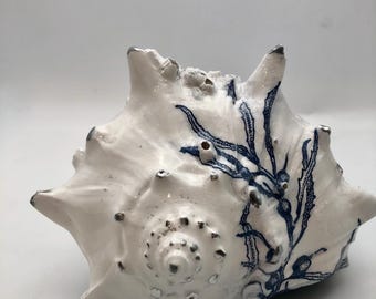 May include: A white seashell featuring a detailed blue botanical illustration of seaweed. The shell has a glossy finish with prominent spikes and a spiral center, accented with metallic silver tips on the points and small circular details.