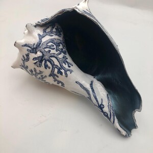 K&ouml;nnte beinhalten: Eine gro&szlig;e Muschelschale mit wei&szlig;er Au&szlig;enseite, die mit komplizierten blauen Korallenmustern verziert ist. Das Innere der Muschel ist in einem tiefen Dunkelblau gehalten, was einen starken Kontrast zwischen der Au&szlig;enfl&auml;che und dem Hohlraum erzeugt.