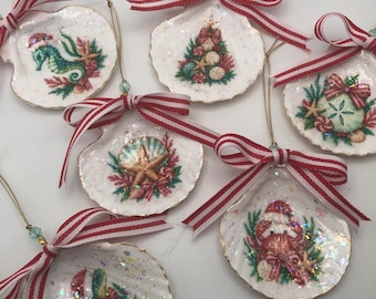 May include: Six white seashell ornaments featuring festive coastal illustrations including a seahorse, a crab, a sea turtle, and holiday motifs. Each shell has a gold-trimmed edge, a shimmering iridescent finish, and a red and white striped ribbon bow.