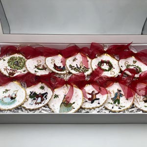 Könnte beinhalten: Ein Set aus zwölf dekorativen Austernschalen-Anhängern in einer weißen Schachtel. Jede Schale zeigt ein handgemaltes Motiv der zwölf Weihnachtstage, goldene Ränder und eine Schleife aus durchscheinendem dunkelrotem Band.