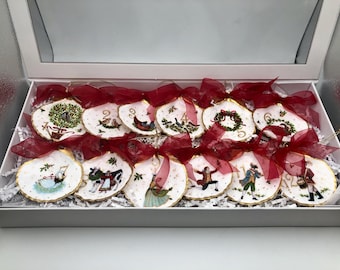 May include: A set of twelve decorative oyster shell ornaments arranged in a white box. Each shell features a unique hand-painted scene from the Twelve Days of Christmas, gold-painted edges, and a translucent deep red ribbon bow.