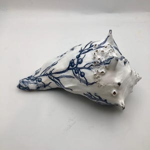 K&ouml;nnte beinhalten: Eine gro&szlig;e wei&szlig;e Muschel mit einer detaillierten blauen botanischen Illustration von Zweigen mit Beeren. Die Muschel hat eine strukturierte Oberfl&auml;che mit nat&uuml;rlichen Graten, spitzen Vorspr&uuml;ngen und kleinen pockenartigen Formationen.
