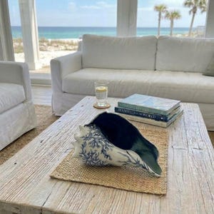 K&ouml;nnte beinhalten: Eine gro&szlig;e Muschel mit wei&szlig;er Au&szlig;enseite, blauem Korallenmuster und dunkelblauem Inneren. Die Muschel liegt auf einer geflochtenen Matte neben zwei B&uuml;chern mit den Titeln Coastal Living und Seashells of the World auf einem Holztisch.