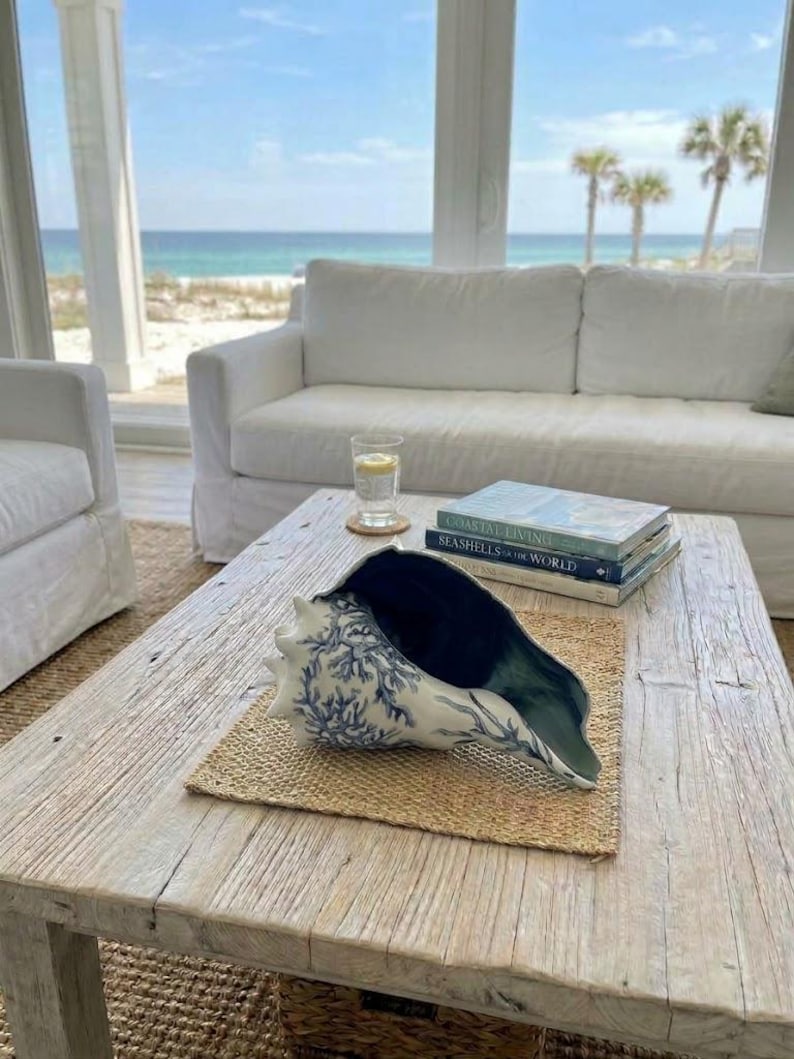 K&ouml;nnte beinhalten: Eine gro&szlig;e Muschel mit wei&szlig;er Au&szlig;enseite, blauem Korallenmuster und dunkelblauem Inneren. Die Muschel liegt auf einer geflochtenen Matte neben zwei B&uuml;chern mit den Titeln Coastal Living und Seashells of the World auf einem Holztisch.