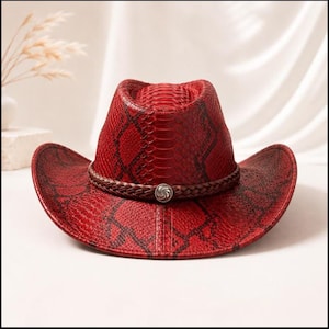 Peut inclure: Un chapeau de cowboy rouge avec un motif peau de serpent noir sur la calotte et les bords. Le chapeau est orné d'une bande tressée marron foncé qui entoure la base de la calotte et comporte un bouton métallique argenté au centre.