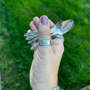 May include: A silver-toned metal ring crafted from a repurposed spoon handle, featuring an embossed floral pattern on the band. The ring is worn on a thumb, while several matching silver-toned spoons are held in the same hand.