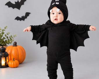 May include: A black long-sleeved hooded jumpsuit featuring bat wings attached to the sides. The hood includes two black ears, white oval eyes, and two white fangs hanging from the top edge, creating a bat-themed costume for an infant.