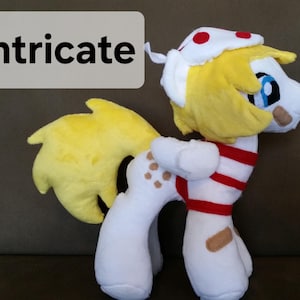 Pode incluir: Um cavalo de pel&uacute;cia branco com uma camisa listrada vermelha e branca, um chap&eacute;u branco com bolinhas vermelhas e uma juba e cauda amarelas. O cavalo tem manchas marrons no corpo e uma bandagem na perna.