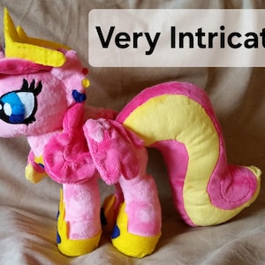 Pode incluir: Brinquedo de pel&uacute;cia rosa e amarelo com uma coroa, asas e uma cauda longa. O brinquedo tem um rosto detalhado com olhos azuis e um nariz rosa.