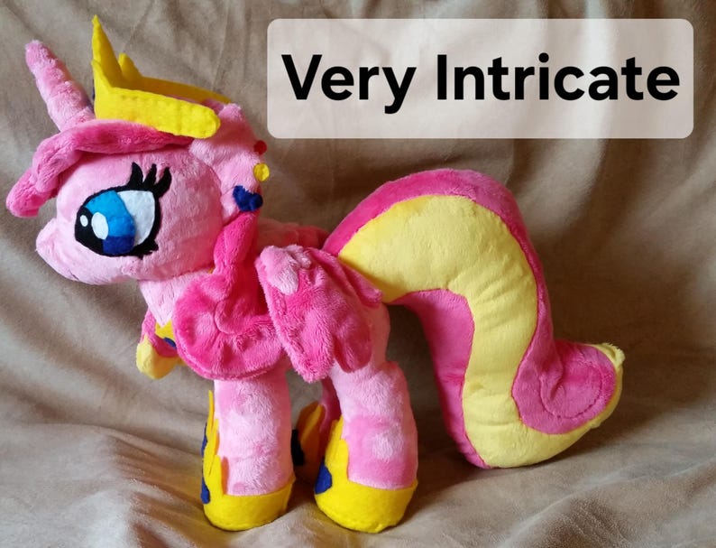 Pode incluir: Brinquedo de pel&uacute;cia rosa e amarelo com uma coroa, asas e uma cauda longa. O brinquedo tem um rosto detalhado com olhos azuis e um nariz rosa.