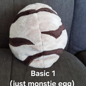 May include: A white and brown striped plush egg. The egg is sitting on a gray couch. The text "Basic 1 (just monstie egg)" is below the egg.