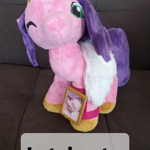 Pode incluir: Unic&oacute;rnio de pel&uacute;cia rosa com cabelo roxo e acabamento de pele branca. O unic&oacute;rnio est&aacute; usando uma coroa dourada e tem uma pequena moldura de foto presa ao seu lado.