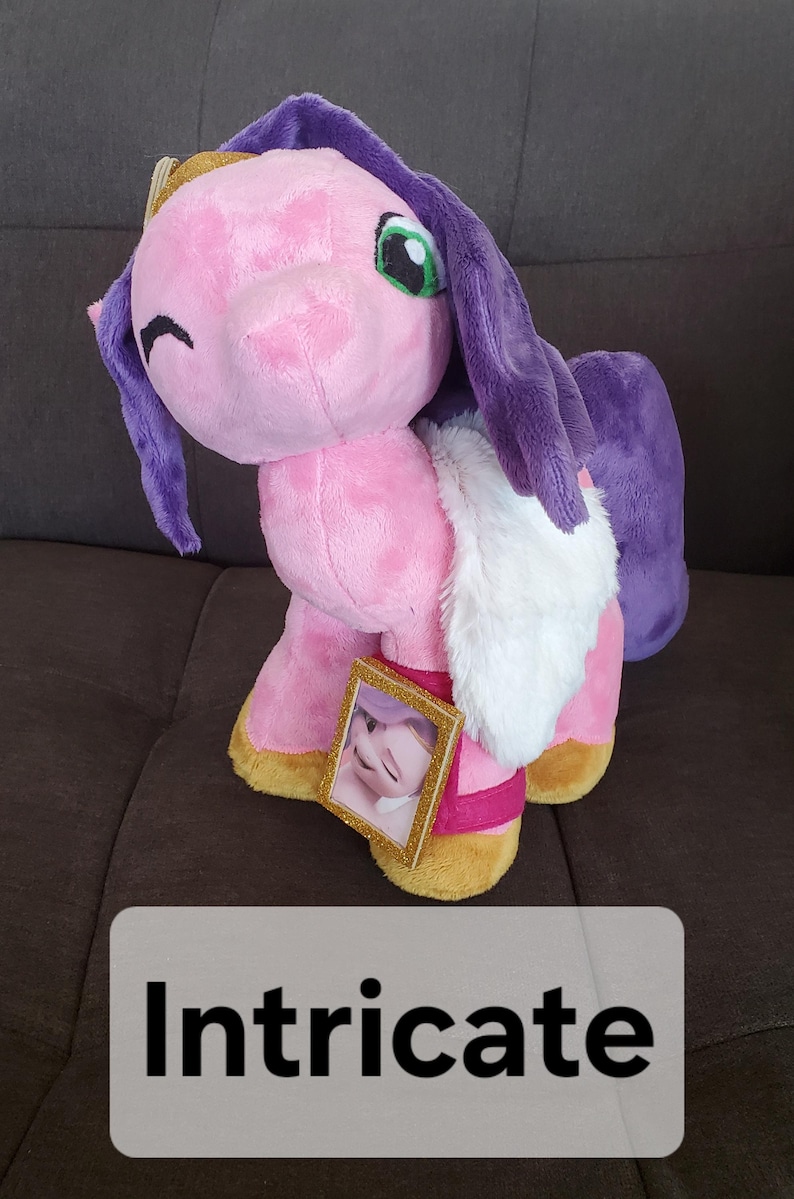 Pode incluir: Unic&oacute;rnio de pel&uacute;cia rosa com cabelo roxo e acabamento de pele branca. O unic&oacute;rnio est&aacute; usando uma coroa dourada e tem uma pequena moldura de foto presa ao seu lado.