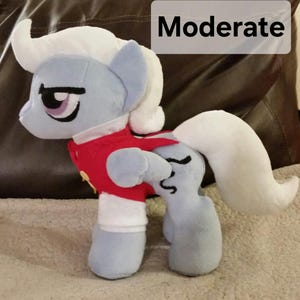 Pode incluir: Um p&ocirc;nei de pel&uacute;cia cinza e branco com um colete vermelho e uma camisa branca. O p&ocirc;nei tem uma crina e uma cauda brancas. O p&ocirc;nei est&aacute; em p&eacute; sobre uma superf&iacute;cie marrom. A palavra "Moderado" est&aacute; escrita em branco sobre um fundo preto.