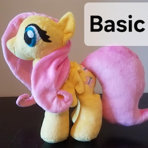 Pode incluir: Um brinquedo de pel&uacute;cia da Pinkie Pie de My Little Pony. O brinquedo &eacute; amarelo com uma juba e uma cauda cor-de-rosa. O brinquedo est&aacute; em p&eacute; sobre uma superf&iacute;cie de madeira. A palavra "Basic" est&aacute; escrita em um ret&acirc;ngulo branco atr&aacute;s do brinquedo.