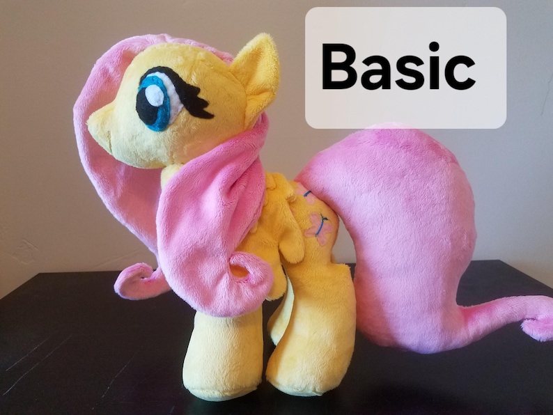 Pode incluir: Um brinquedo de pel&uacute;cia da Pinkie Pie de My Little Pony. O brinquedo &eacute; amarelo com uma juba e uma cauda cor-de-rosa. O brinquedo est&aacute; em p&eacute; sobre uma superf&iacute;cie de madeira. A palavra "Basic" est&aacute; escrita em um ret&acirc;ngulo branco atr&aacute;s do brinquedo.