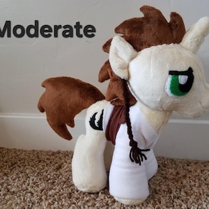 Pode incluir: Um cavalo de pel&uacute;cia com pelo marrom e branco, uma camisa branca e uma tran&ccedil;a marrom. O cavalo tem olhos verdes e um s&iacute;mbolo de lua crescente preto no seu lado. A palavra "Moderado" est&aacute; escrita em texto preto sobre um fundo branco.