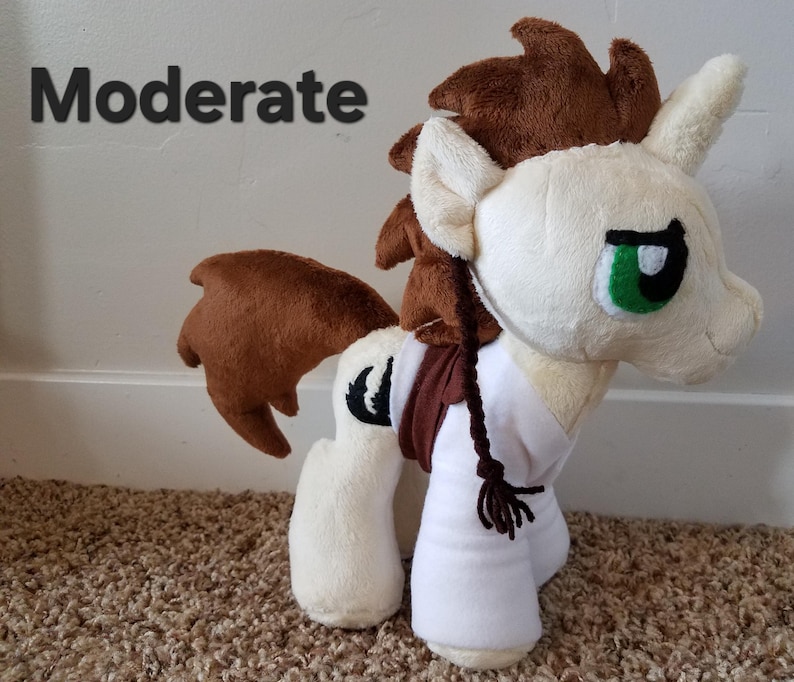 Pode incluir: Um cavalo de pel&uacute;cia com pelo marrom e branco, uma camisa branca e uma tran&ccedil;a marrom. O cavalo tem olhos verdes e um s&iacute;mbolo de lua crescente preto no seu lado. A palavra "Moderado" est&aacute; escrita em texto preto sobre um fundo branco.