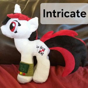 Pode incluir: Unic&oacute;rnio de pel&uacute;cia branco com crina e cauda vermelhas e pretas. O unic&oacute;rnio tem um olho vermelho, um nariz preto e uma boca preta. O unic&oacute;rnio tem um s&iacute;mbolo de naipe de cartas preto e vermelho no seu lado. O unic&oacute;rnio est&aacute; usando uma pulseira verde e amarela com o texto "D.S.F.N.L." nela.